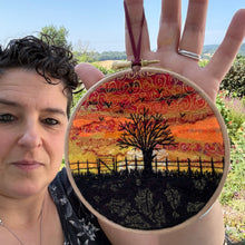 A person outdoors holds up a round embroidery hoop featuring a colorful embroidered landscape with a tree, fence, and swirling sky pattern in red, orange, yellow, black, and green.